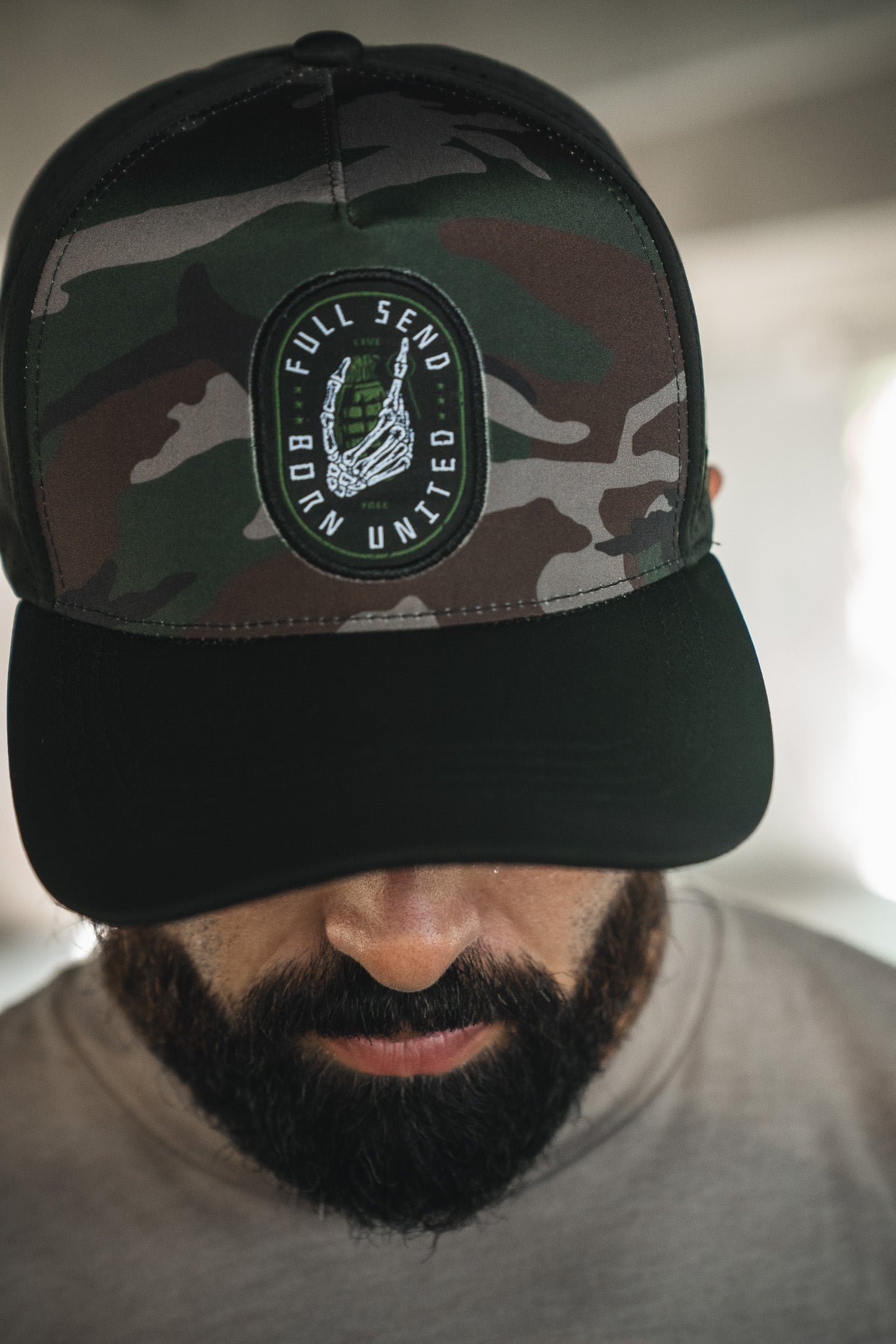 A bearded man wears the Born United Premium Snapback - Full Send Camo by Born United Private Label, featuring a camo design and "FULL SEND BORN UNITED" patch, as he looks down with a softly blurred background.