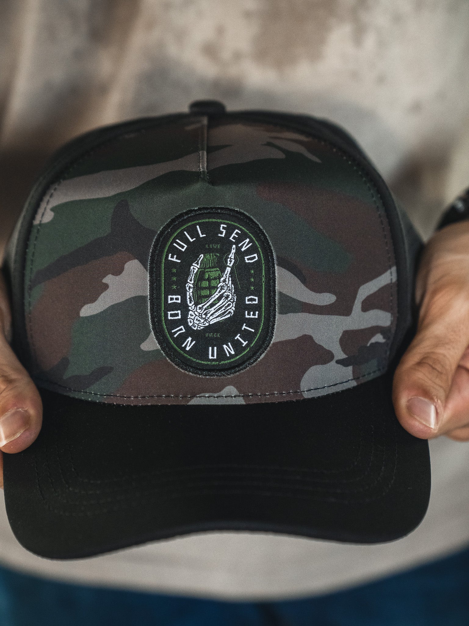 A person holds the Born United Private Label Born United Premium Snapback - Full Send (Camo), a camouflage-patterned cap with a black brim and a patch featuring "FULL SEND UNITED" around a helmet and laurel wreath.