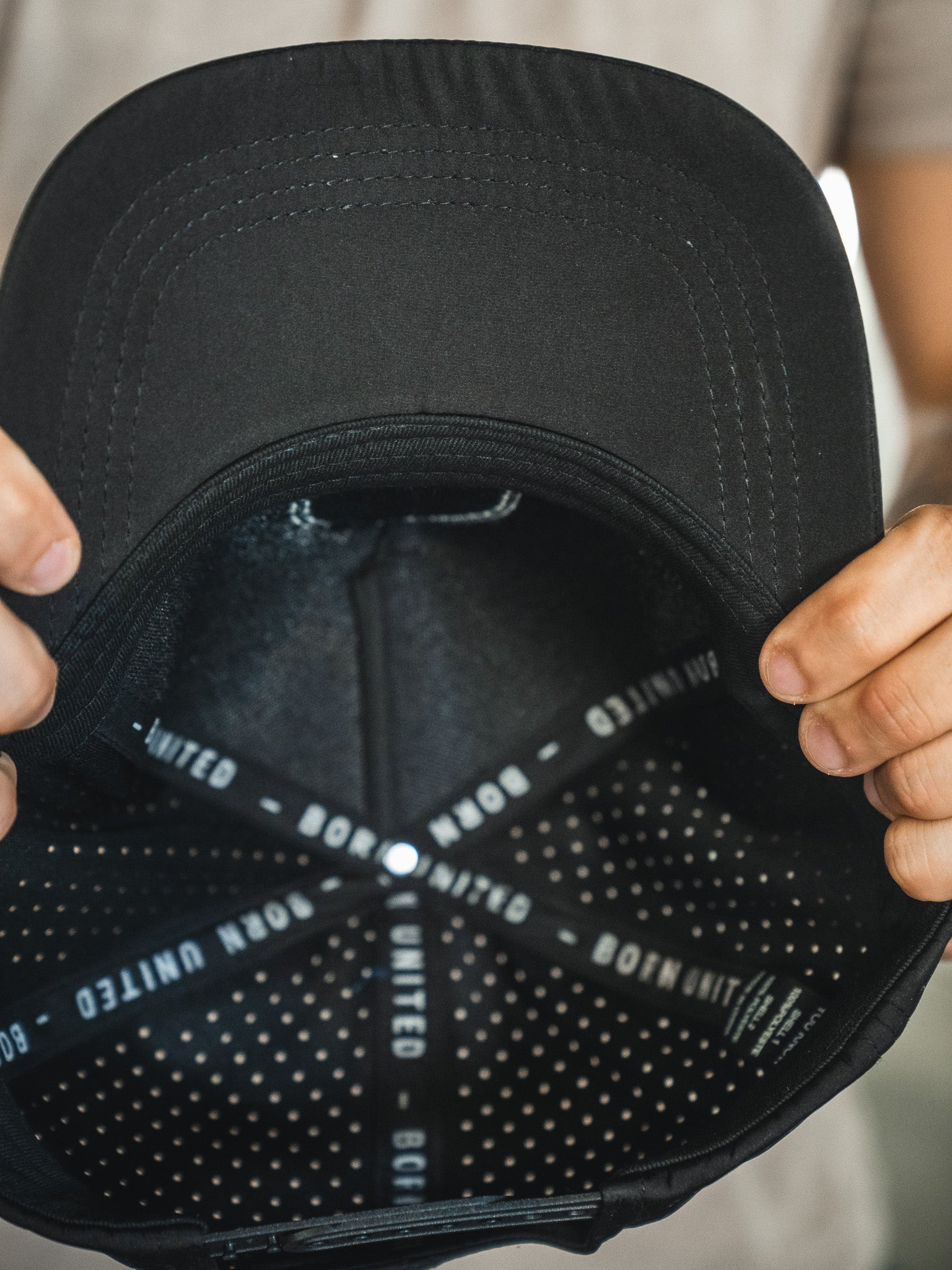 A person holds the Born United Premium Snapback - Full Send (Camo) by Born United Private Label inside-out, showing the "BORN UNITED" text on the inner band and a dotted interior pattern. Only their hands are visible.
