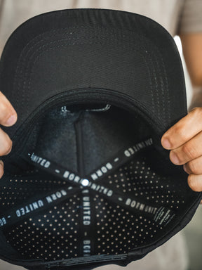 A person holds the Born United Premium Snapback - Full Send (Camo) by Born United Private Label inside-out, showing the "BORN UNITED" text on the inner band and a dotted interior pattern. Only their hands are visible.