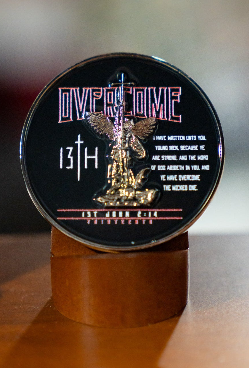 The 13th Overcome Challenge Coin is round and upright, showing an angel defeating a dragon, with “OVERCOME,” “13th,” and a biblical quote from 1 John 2:14 in white text on a dark background.