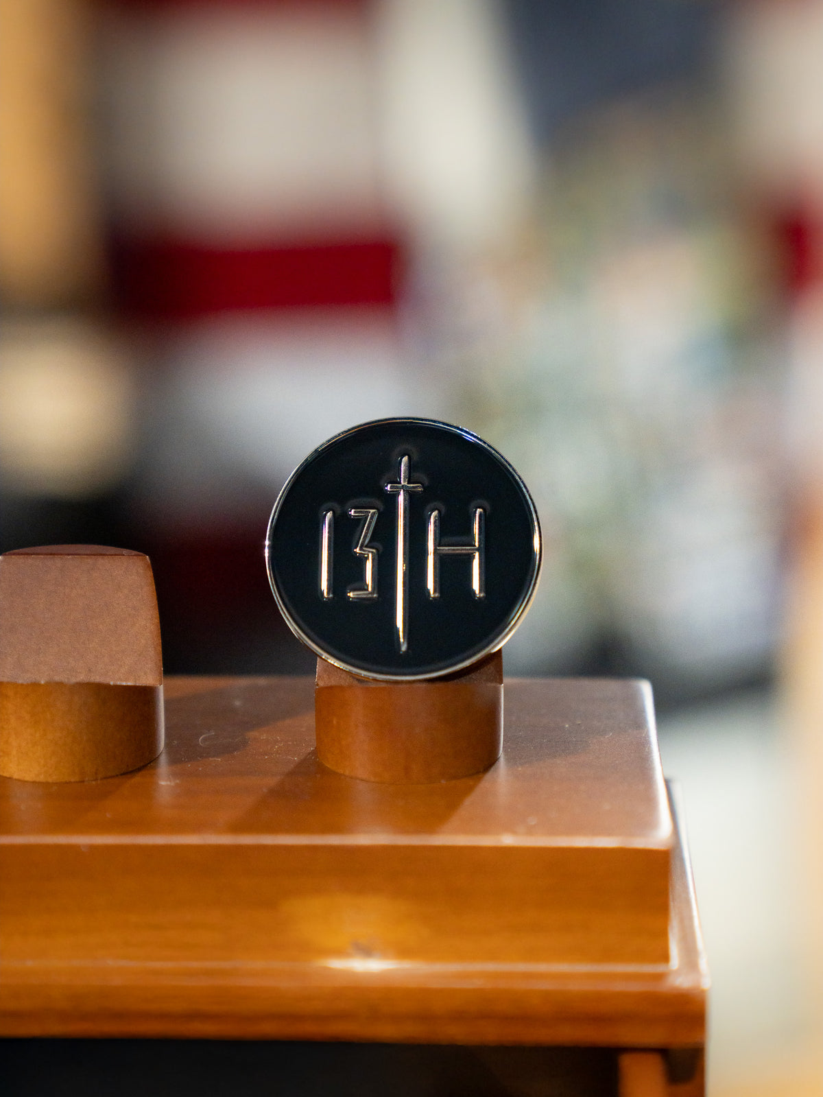 The Overcome Challenge Coin by 13th is a black oval plaque with silver 13TH markings, standing upright on wood, symbolizing resilience amid a blurred, colorful background.