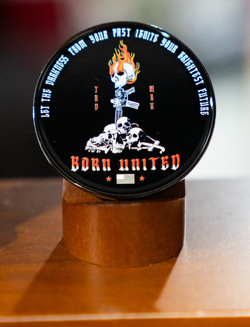 The Let the Darkness Challenge Coin by Born United Private Label features a round black tin with a flaming skull, crossed rifles, and central skulls. The tin reads “LET THE DARKNESS FROM YOUR PAST IGNITE YOUR BRIGHTEST FUTURE” and “BORN UNITED,” plus an American flag.