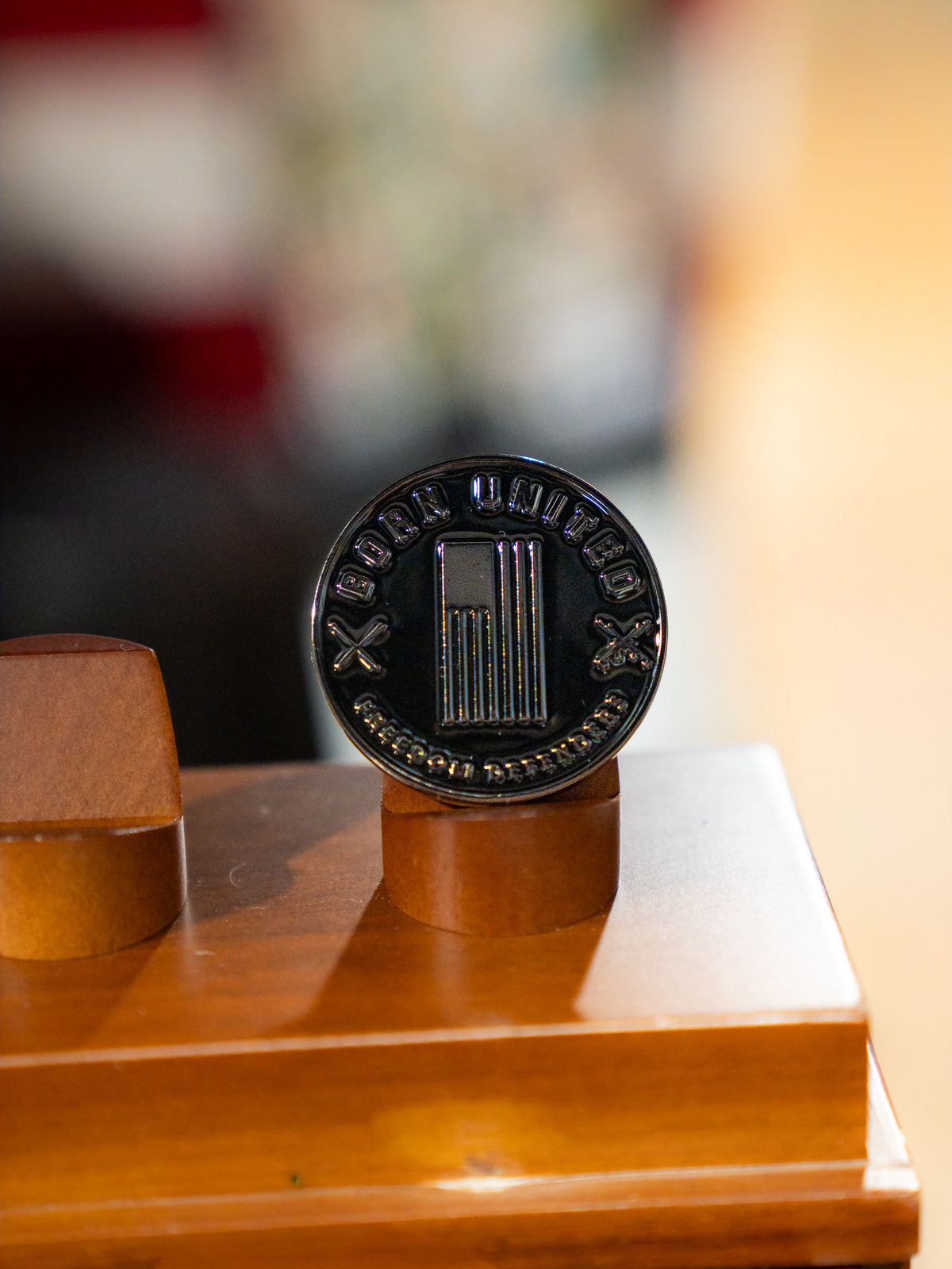 A close-up of the round, black Honey Badgerin' Challenge Coin by Born United Private Label, featuring a pillar design with "BODY UNITED" and "ACCEPT NO DEFEATS" engraved, shown on a wooden stand. Ideal for collectors and Honey Badger fans.