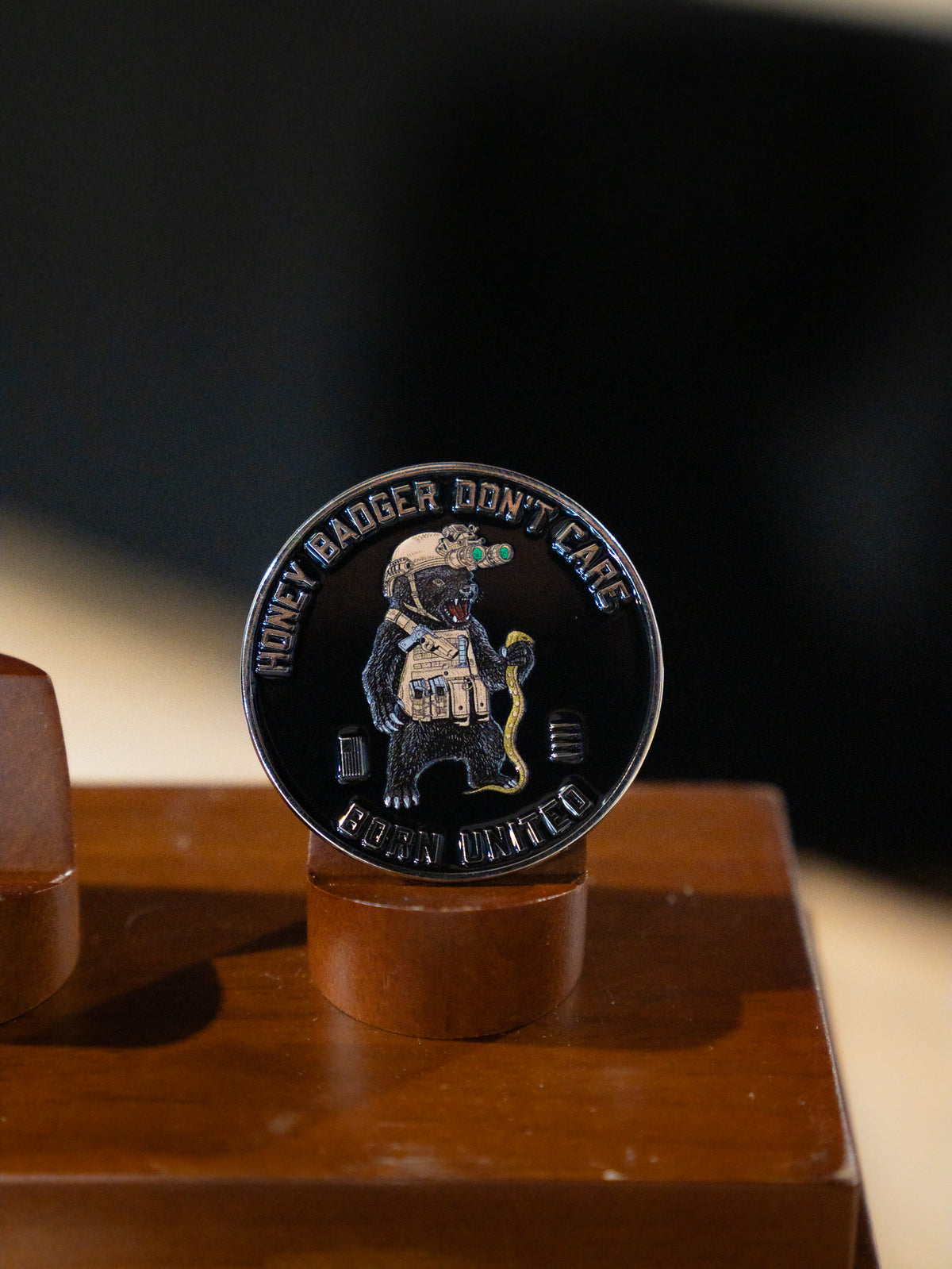 The Honey Badgerin' Challenge Coin by Born United Private Label features an armored honey badger with a snake and night-vision goggles, plus "HONEY BADGER DONT CARE" and "CUBAN UNITED" text around the edge, shown on a wooden stand.