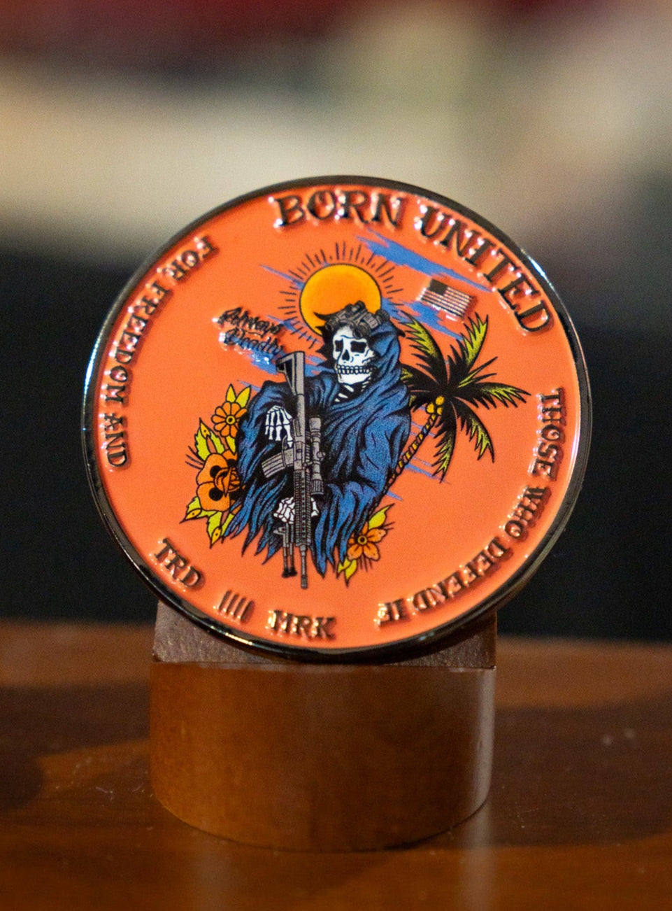 The Always Deadly Challenge Coin by Born United Private Label is a round, orange coin showing a skeleton in a blue cloak with a rifle, plus palm tree, sun, and smoke. The edge reads: "BORN UNITED, FOR THOSE OF KIND, FOR THOSE OF DEVASTATION—Always Deadly.