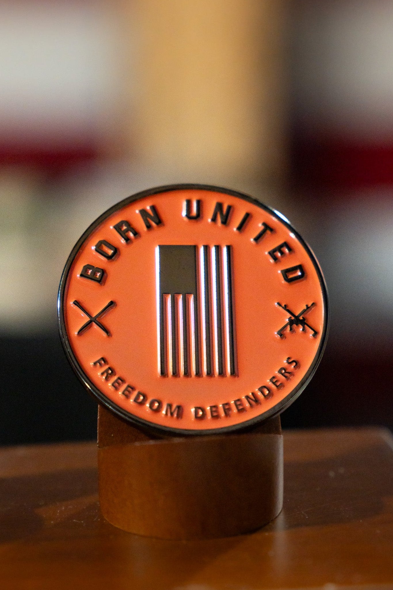 The Born United Private Label "Always Deadly Challenge Coin" features an American flag, “BORN UNITED” on top, “FREEDOM DEFENDERS” below, two crossed rifles, and the phrase Always Deadly. Displayed on a wooden stand.