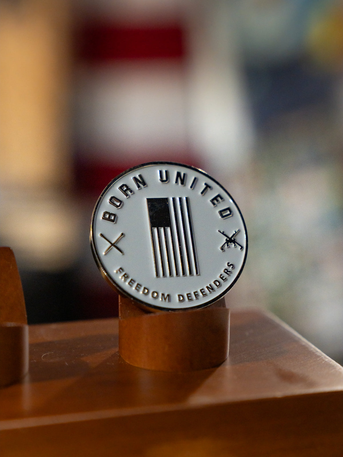 The Red Line Flag Challenge Coin by Born United Private Label features a stylized American flag, crossed rifles, and the words BORN UNITED and FREEDOM DEFENDERS—ideal fan gear displayed on a wooden stand.