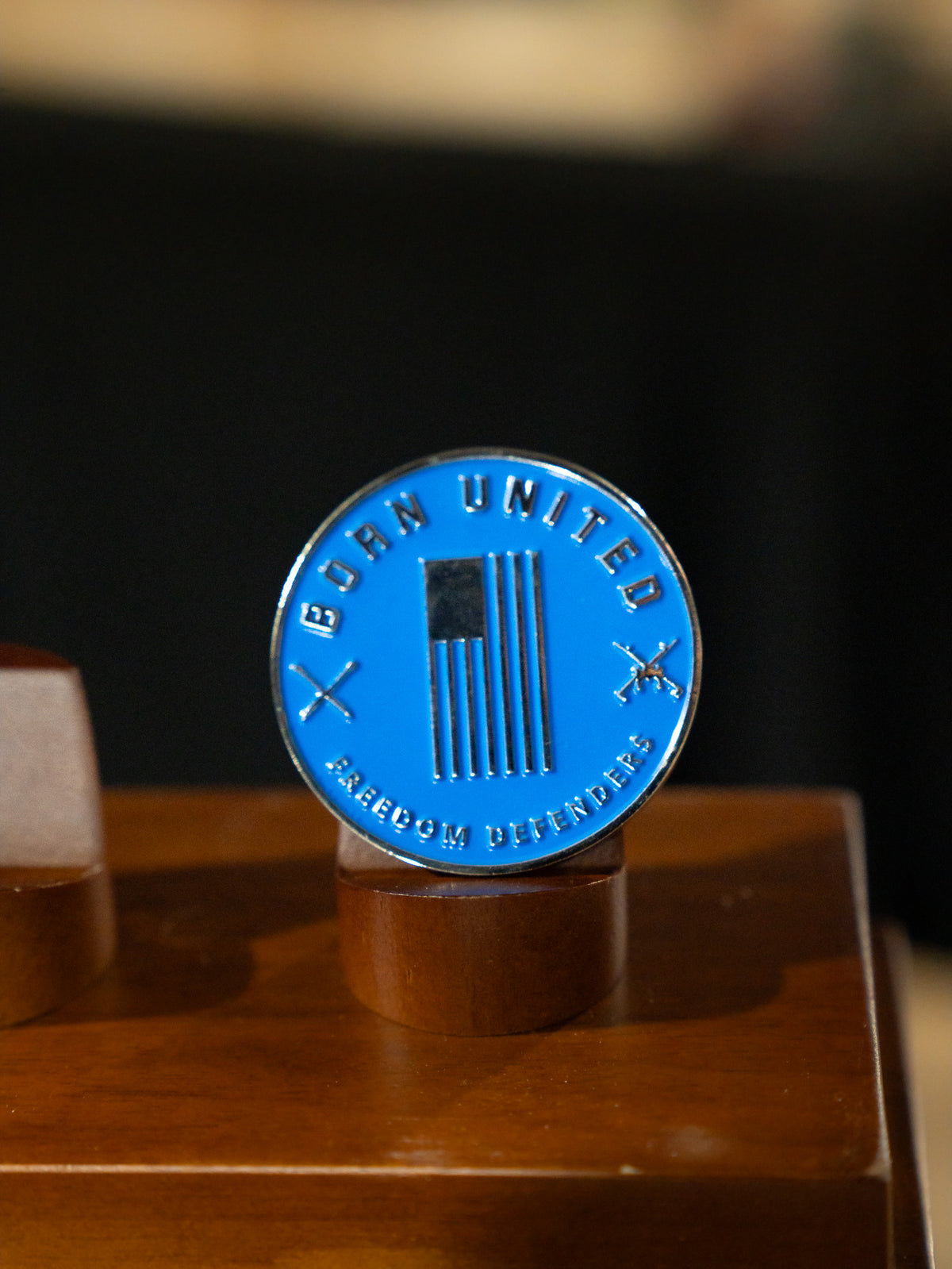 The Disabled Vet Challenge Coin by Born United Private Label features a black and white American flag, crossed rifles, and the words "Born United" and "Freedom Defender," honoring every disabled vet. Displayed proudly on a wooden stand.