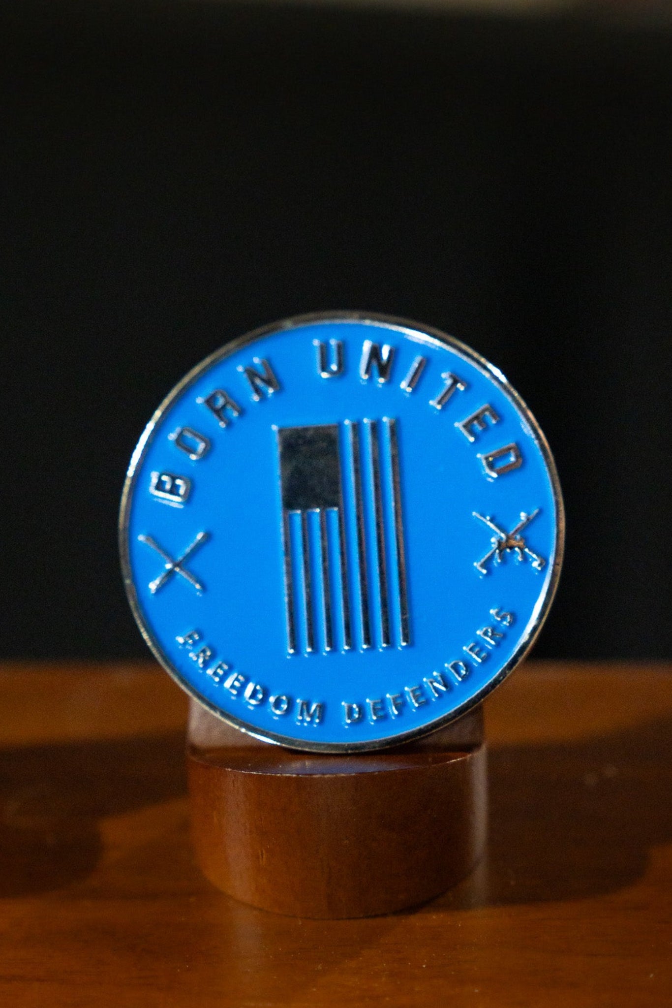The Disabled Vet Challenge Coin by Born United Private Label features a black and white American flag, crossed rifles, and the words "Born United" and "Freedom Defender," honoring every disabled vet. Displayed proudly on a wooden stand.