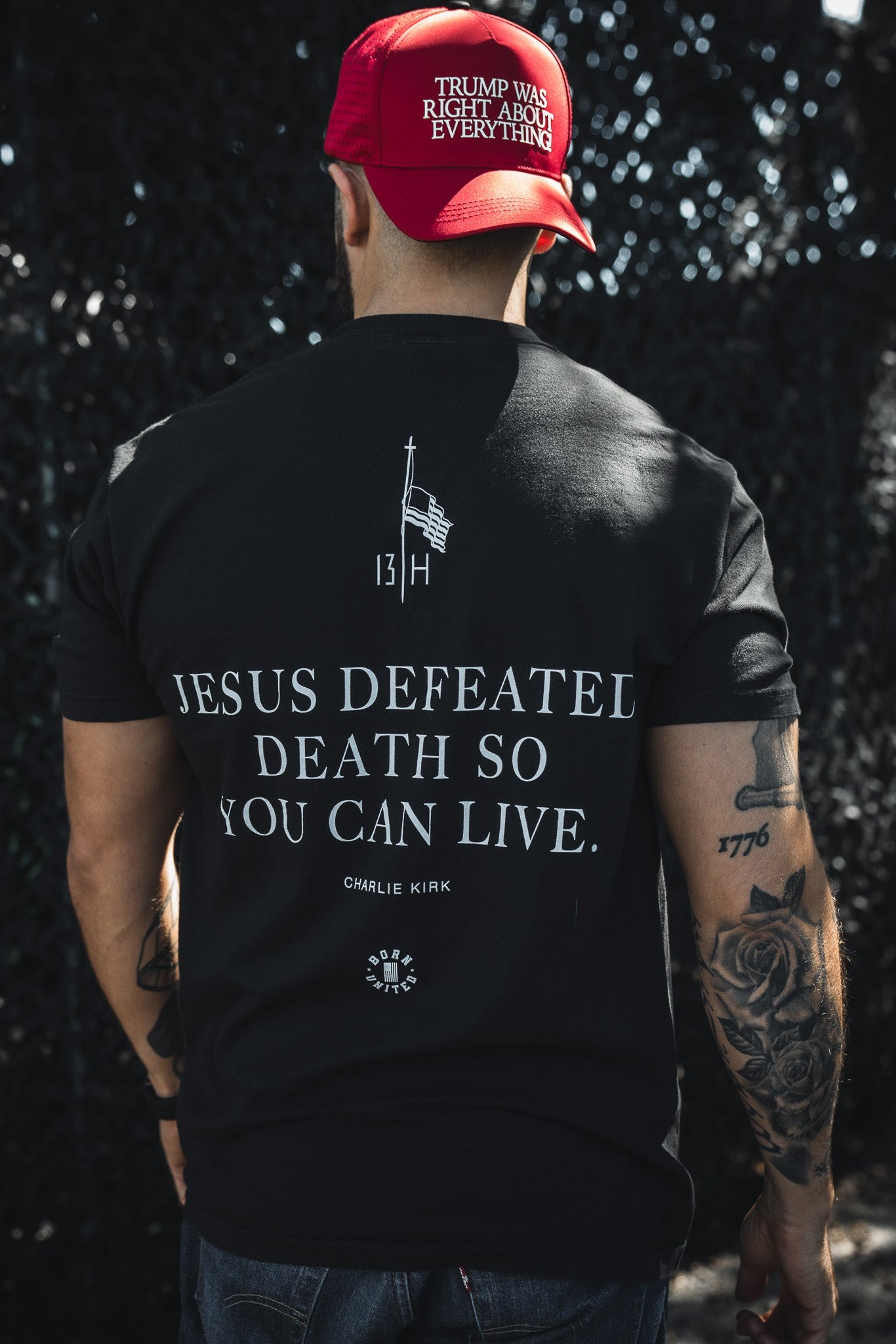 A man with tattooed arms faces away from the camera, wearing a red TRUMP WAS RIGHT ABOUT EVERYTHING cap and a Born United Private Label "Live Like Charlie" t-shirt that reads JESUS DEFEATED DEATH SO YOU CAN LIVE. CHARLIE KIRK.