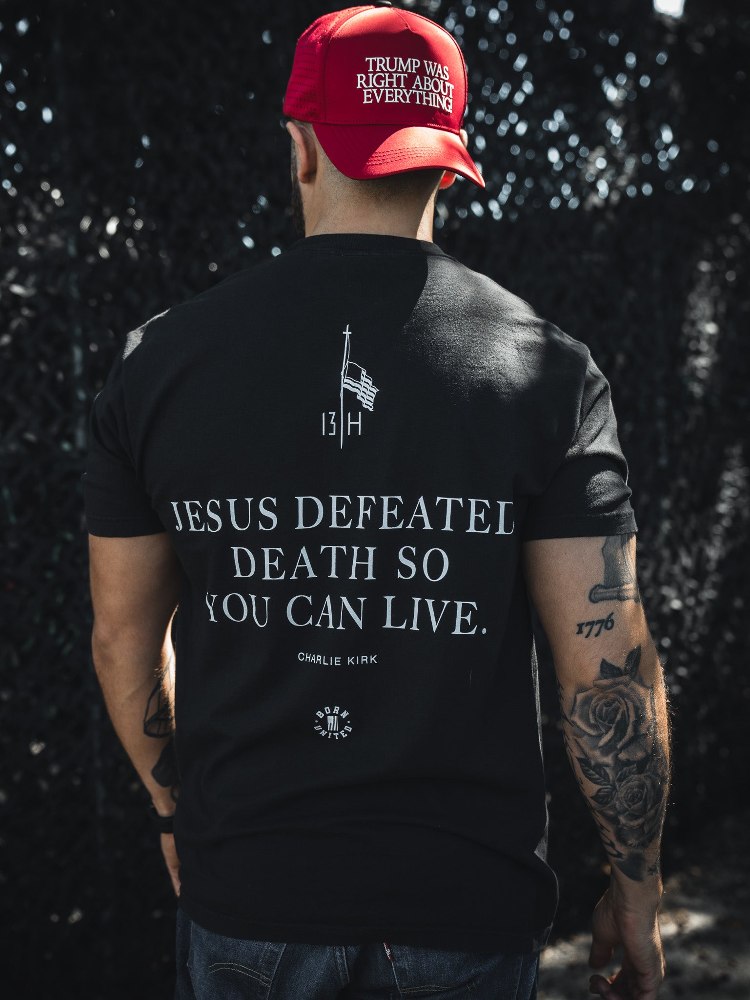 A man with tattooed arms faces away from the camera, wearing a red TRUMP WAS RIGHT ABOUT EVERYTHING cap and a Born United Private Label "Live Like Charlie" t-shirt that reads JESUS DEFEATED DEATH SO YOU CAN LIVE. CHARLIE KIRK.