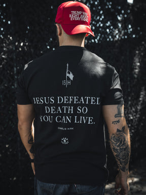 A man with tattooed arms faces away from the camera, wearing a red TRUMP WAS RIGHT ABOUT EVERYTHING cap and a Born United Private Label "Live Like Charlie" t-shirt that reads JESUS DEFEATED DEATH SO YOU CAN LIVE. CHARLIE KIRK.