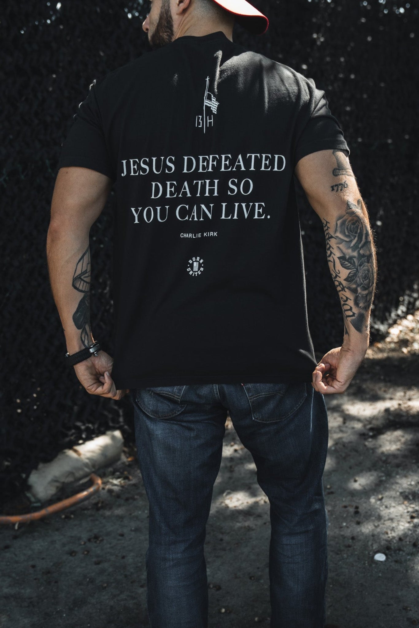 A man stands outdoors with his back to the camera, wearing jeans, a red backwards cap, and a Born United Private Label "Live Like Charlie" t-shirt featuring the message "Jesus defeated death so you can live.