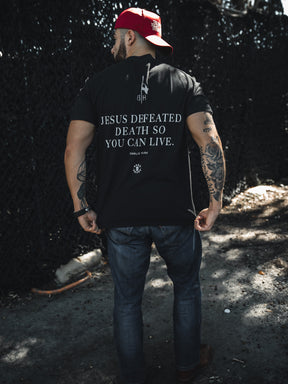 A man stands outdoors with his back to the camera, wearing jeans, a red backwards cap, and a Born United Private Label "Live Like Charlie" t-shirt featuring the message "Jesus defeated death so you can live.