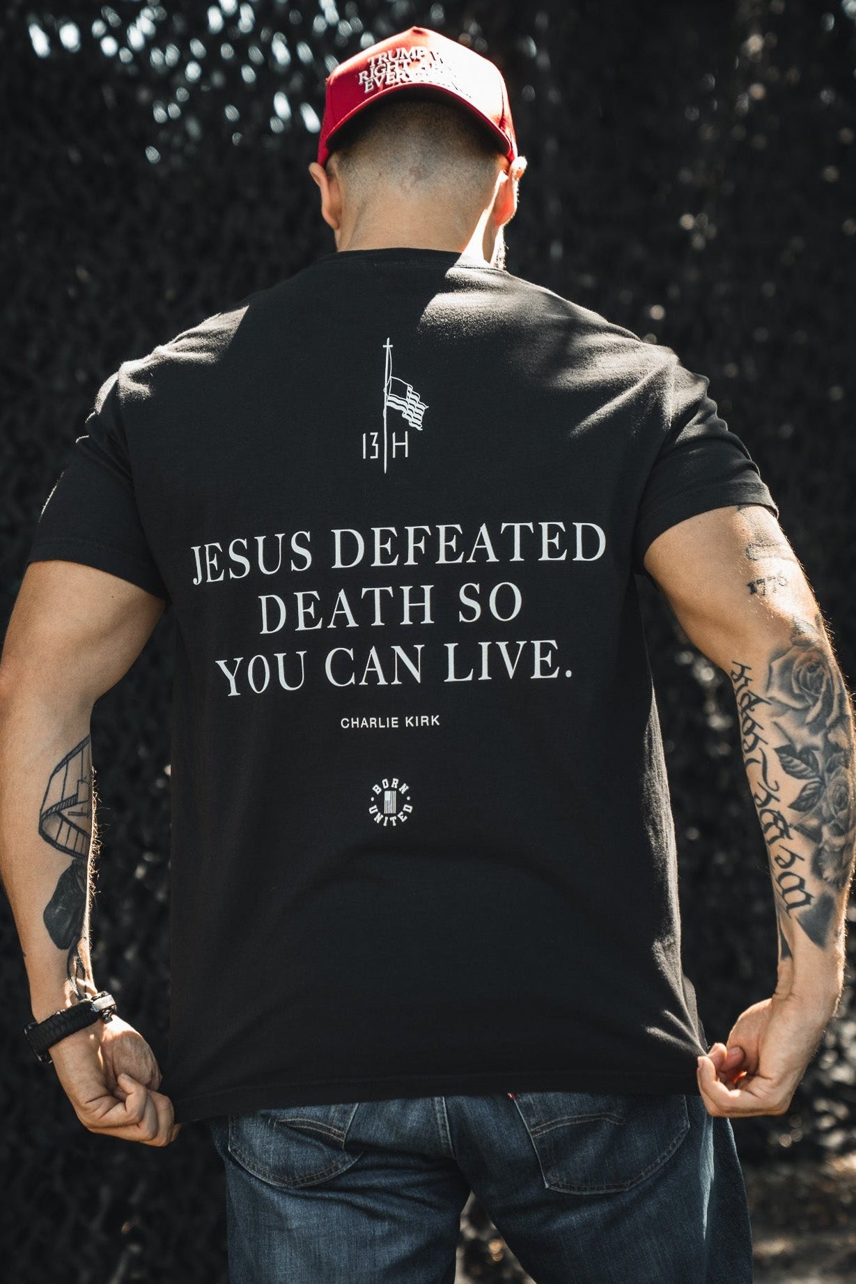 A man in a red cap and black t-shirt faces away, revealing the "Jesus defeated death so you can live" text. The Born United Private Label Live Like Charlie tee features Charlie Kirk’s name, a flag, sun symbol, and highlights Christian values.