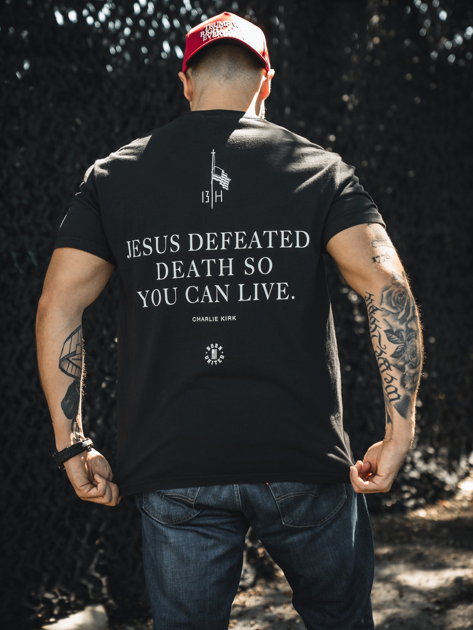 A man in a red cap and black t-shirt faces away, revealing the "Jesus defeated death so you can live" text. The Born United Private Label Live Like Charlie tee features Charlie Kirk’s name, a flag, sun symbol, and highlights Christian values.