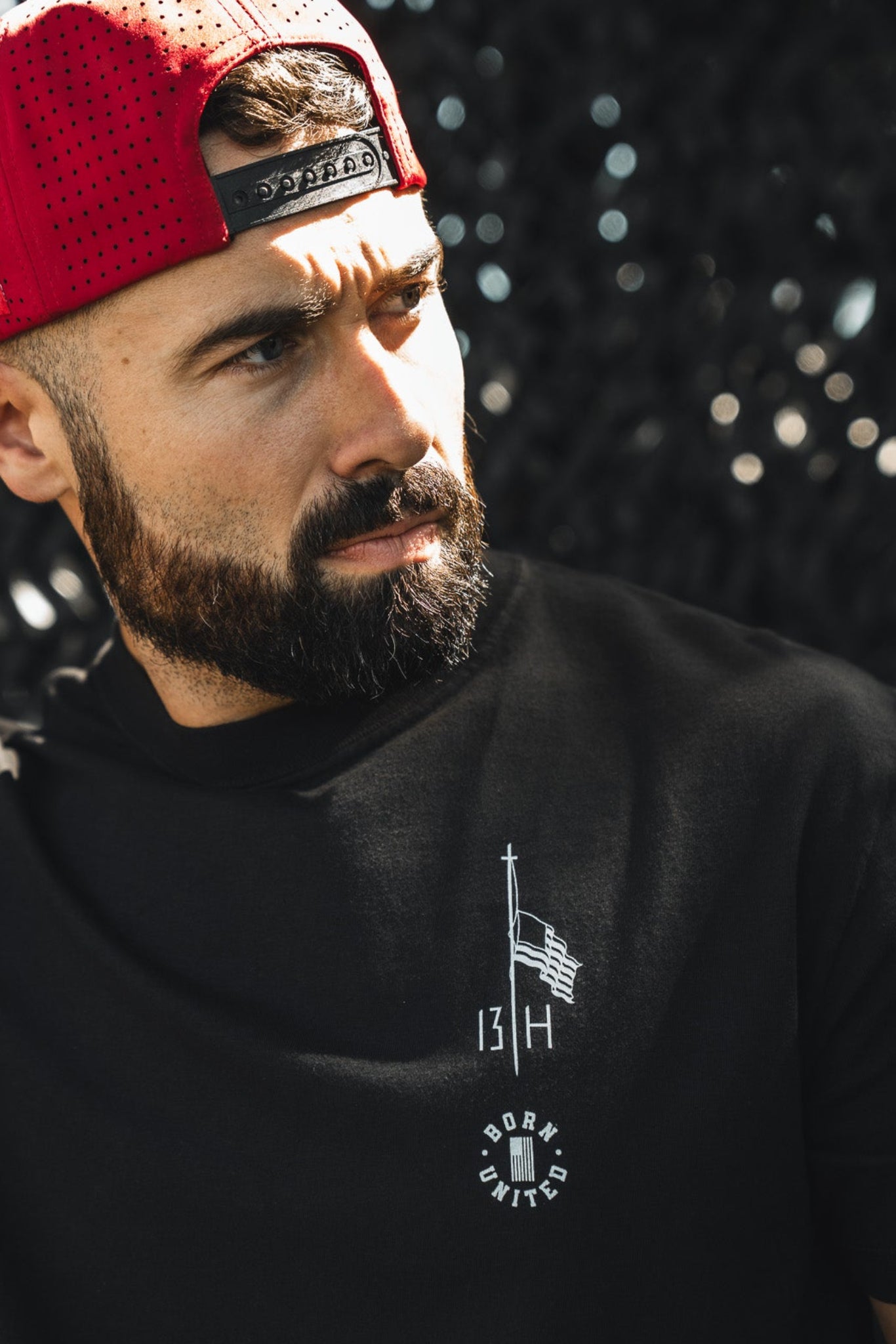 A bearded man in a red backward baseball cap and a Born United Private Label Live Like Charlie tee stands in sunlight, adjusting his hat, with a dark textured background behind him.