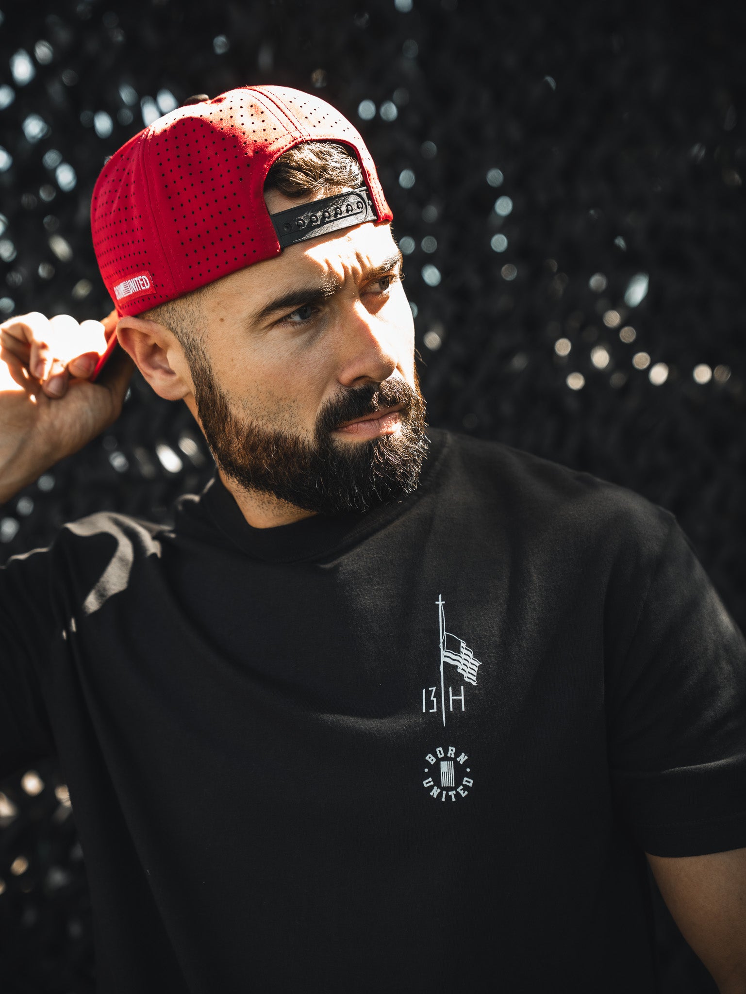 A bearded man in a red backward baseball cap and a Born United Private Label Live Like Charlie tee stands in sunlight, adjusting his hat, with a dark textured background behind him.
