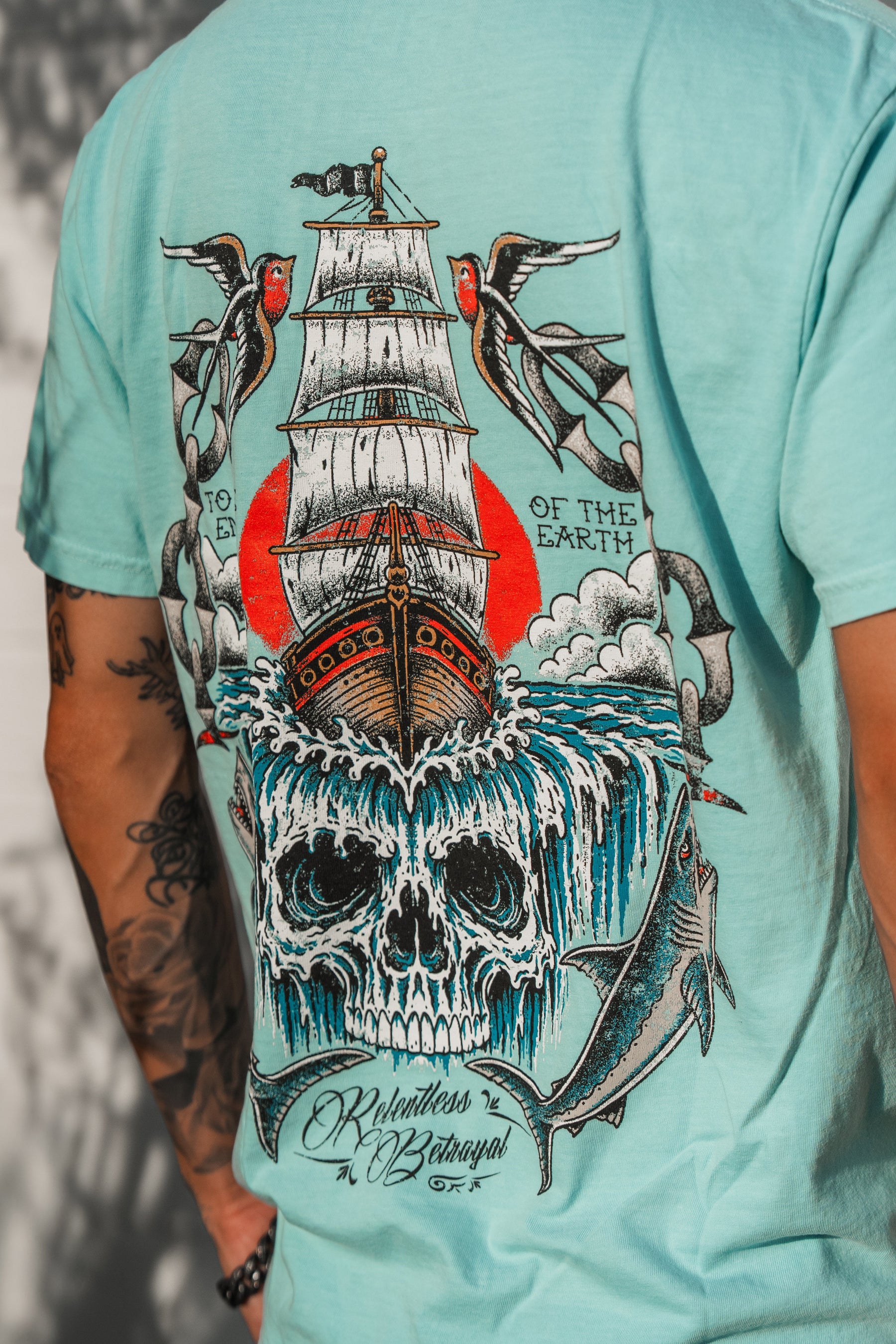 Introducing the To The Ends Of The Earth Premium T-Shirt by Relentless Betrayal—a turquoise unisex shirt featuring a striking graphic of a sailing ship, crashing waves forming a skull, birds, and a shark, along with the text “Relentless as the Ocean, Wild as the Earth.” Crafted from 100% cotton for premium comfort and style.