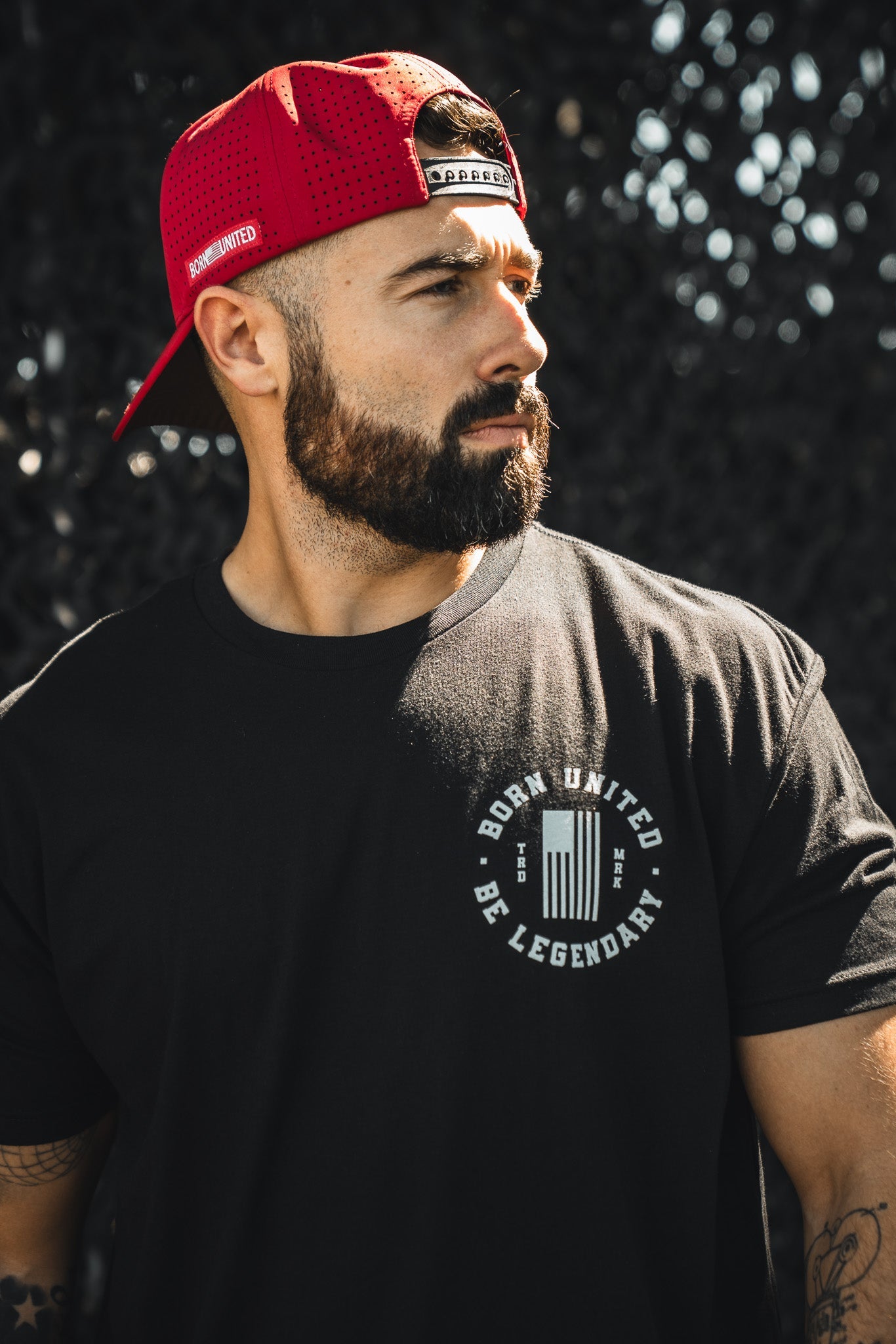 A bearded man in a red Born United Premium Snapback - Trump Was Right cap and a black t-shirt stands outdoors, looking to the side with a serious expression. Brand: Born United Private Label.