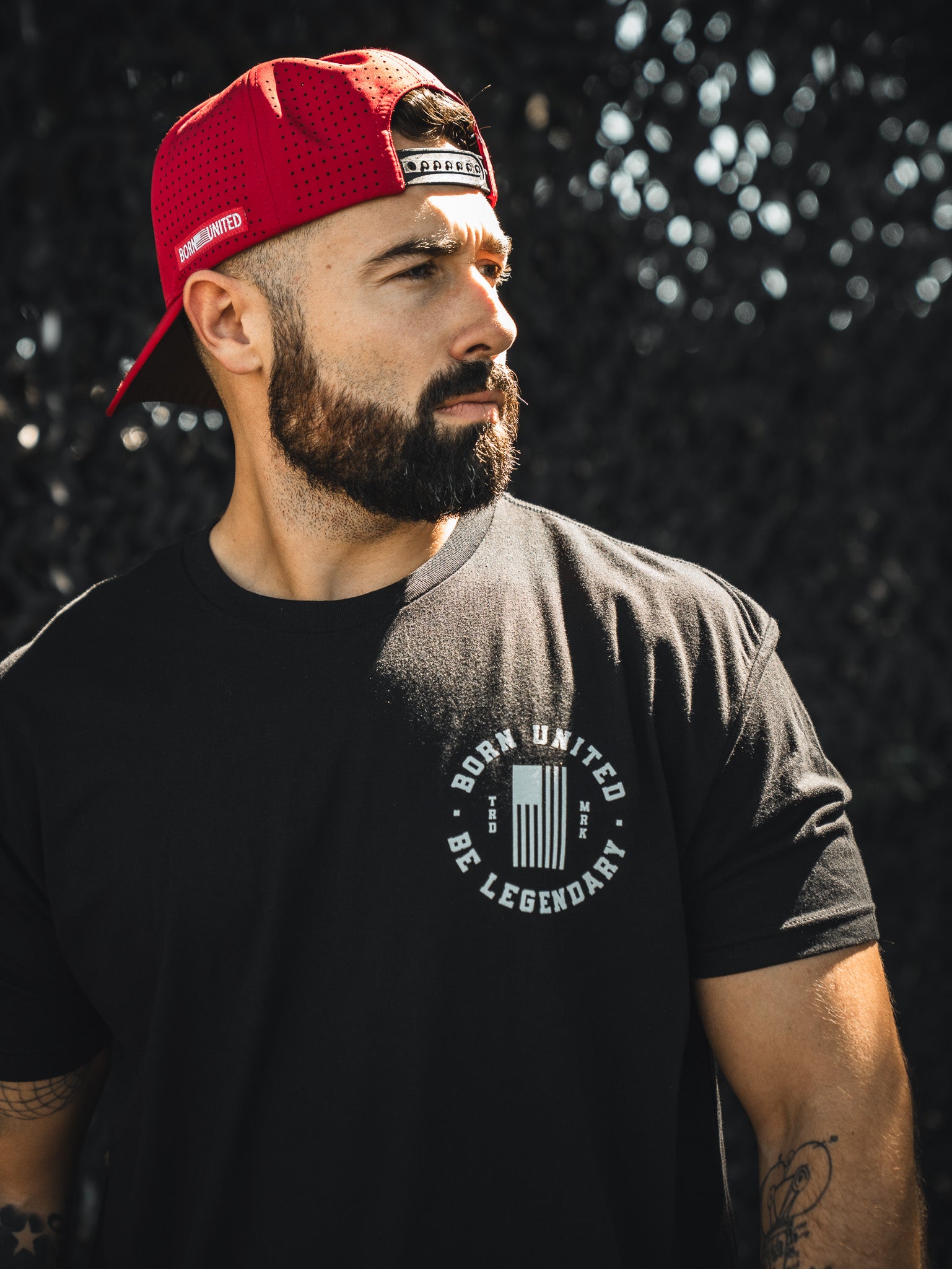A bearded man in a red Born United Premium Snapback - Trump Was Right cap and a black t-shirt stands outdoors, looking to the side with a serious expression. Brand: Born United Private Label.