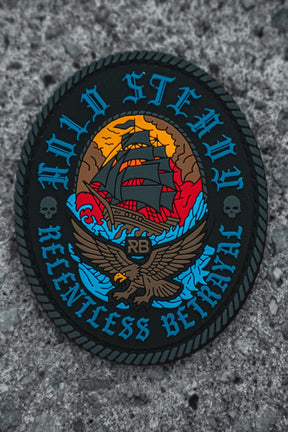 The Hold Steady Patch by Relentless Betrayal is a round 3x3 Velcro patch showing a sailing ship, eagle, skulls, and 'RB' initials on gray. It says “HOLD STEADY” above and “RELENTLESS BETRAYAL” below.
