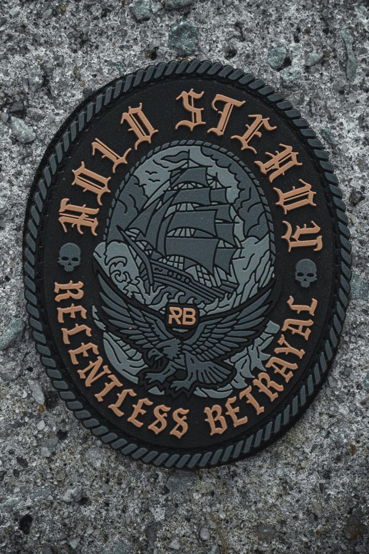 The Hold Steady Ghost Patch by Relentless Betrayal is a 3" embroidered patch with a large ship, eagle, skulls on each side, and bold “HOLD STEADY RELENTLESS BETRAYAL” text. “RB” is centered below the ship. Displayed on rough stone.