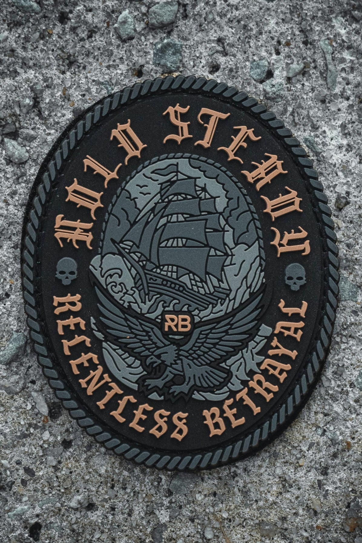 The Hold Steady Ghost Patch by Relentless Betrayal is a black and gray 3" Velcro patch featuring a sailing ship, eagle, skulls, rope border, "HOLD STEADY" on top, "RELENTLESS BETRAYAL" below, and RB in the center. Shown on rough concrete.