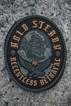 The Hold Steady Ghost Patch by Relentless Betrayal is a black and gray 3" Velcro patch featuring a sailing ship, eagle, skulls, rope border, "HOLD STEADY" on top, "RELENTLESS BETRAYAL" below, and RB in the center. Shown on rough concrete.