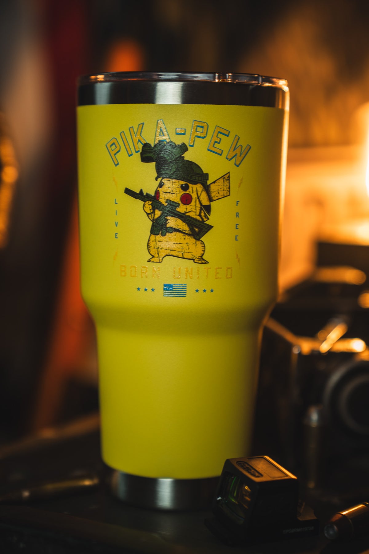 The Born United Private Label Pika-Pew Tumbler - 30oz is a yellow travel tumbler featuring a cartoon Pikachu soldier with a rifle and the text "PIKA-PEW" and "Born United," shown on a table with camera gear nearby.
