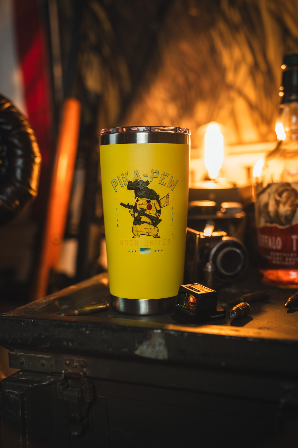The Born United Private Label Pika-Pew Tumbler - 20oz, featuring a yellow design with a PIKA-PEW cartoon and Ukrainian flag, sits on a cluttered table with a camera, bottle, and tools under warm background light.