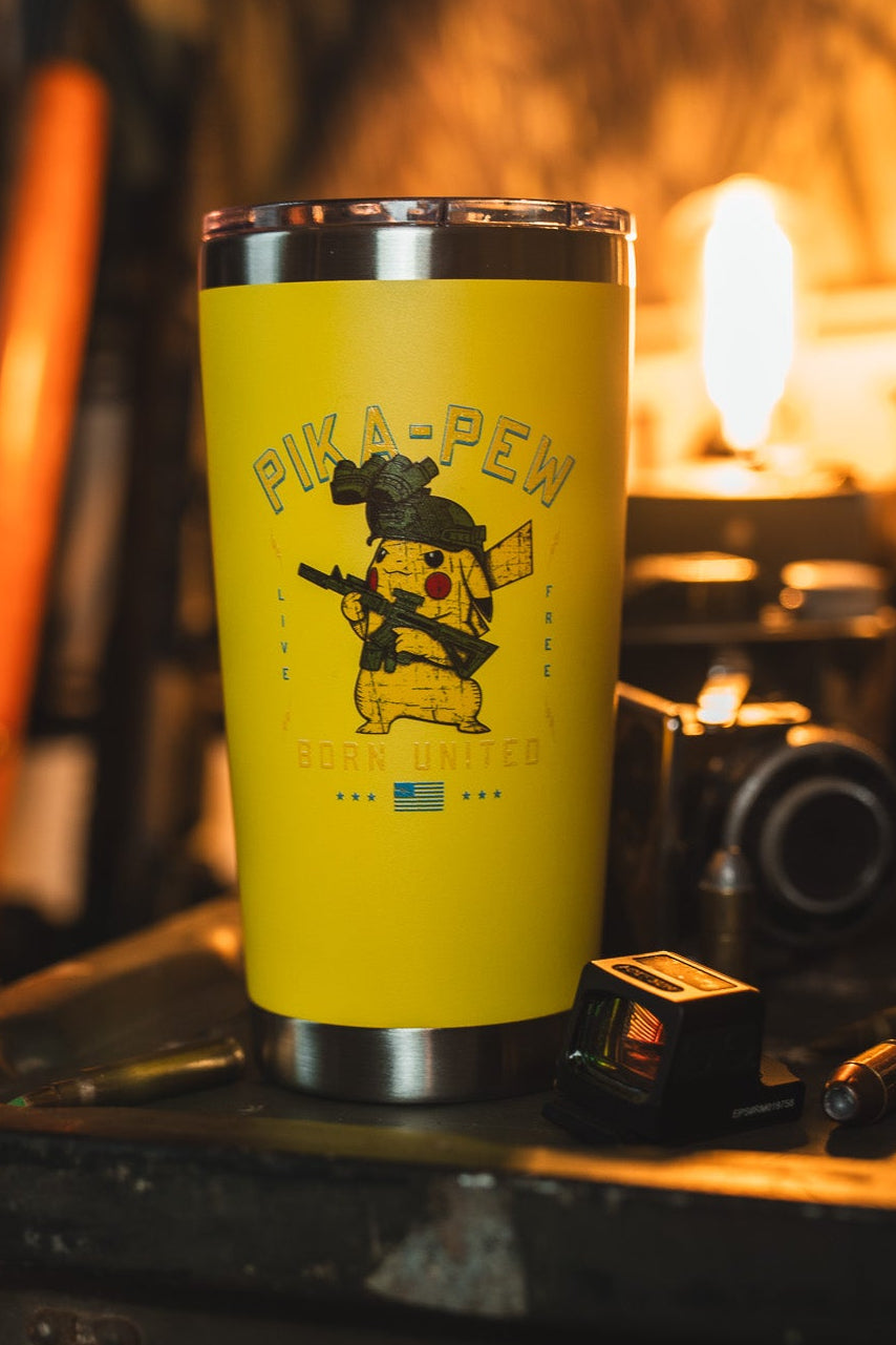 The Born United Private Label Pika-Pew Tumbler - 20oz, featuring a yellow design with a PIKA-PEW cartoon and Ukrainian flag, sits on a cluttered table with a camera, bottle, and tools under warm background light.