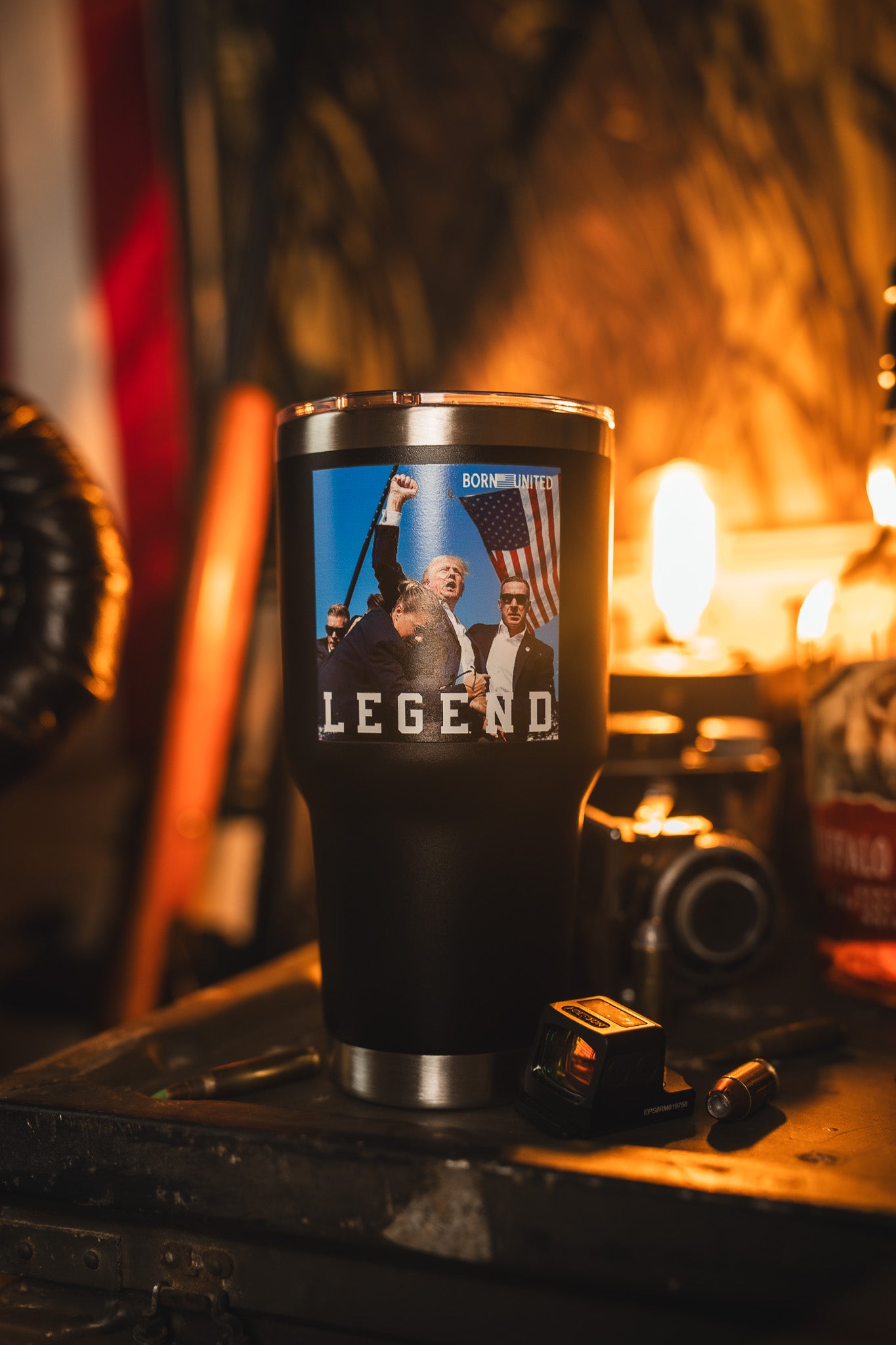 The Born United Private Label Forever Legend Tumbler - 30oz sits on a table, decorated with a sticker of a fist raised next to an American flag and the word LEGEND. Warm lighting and items like a camera and bottle appear in the background.