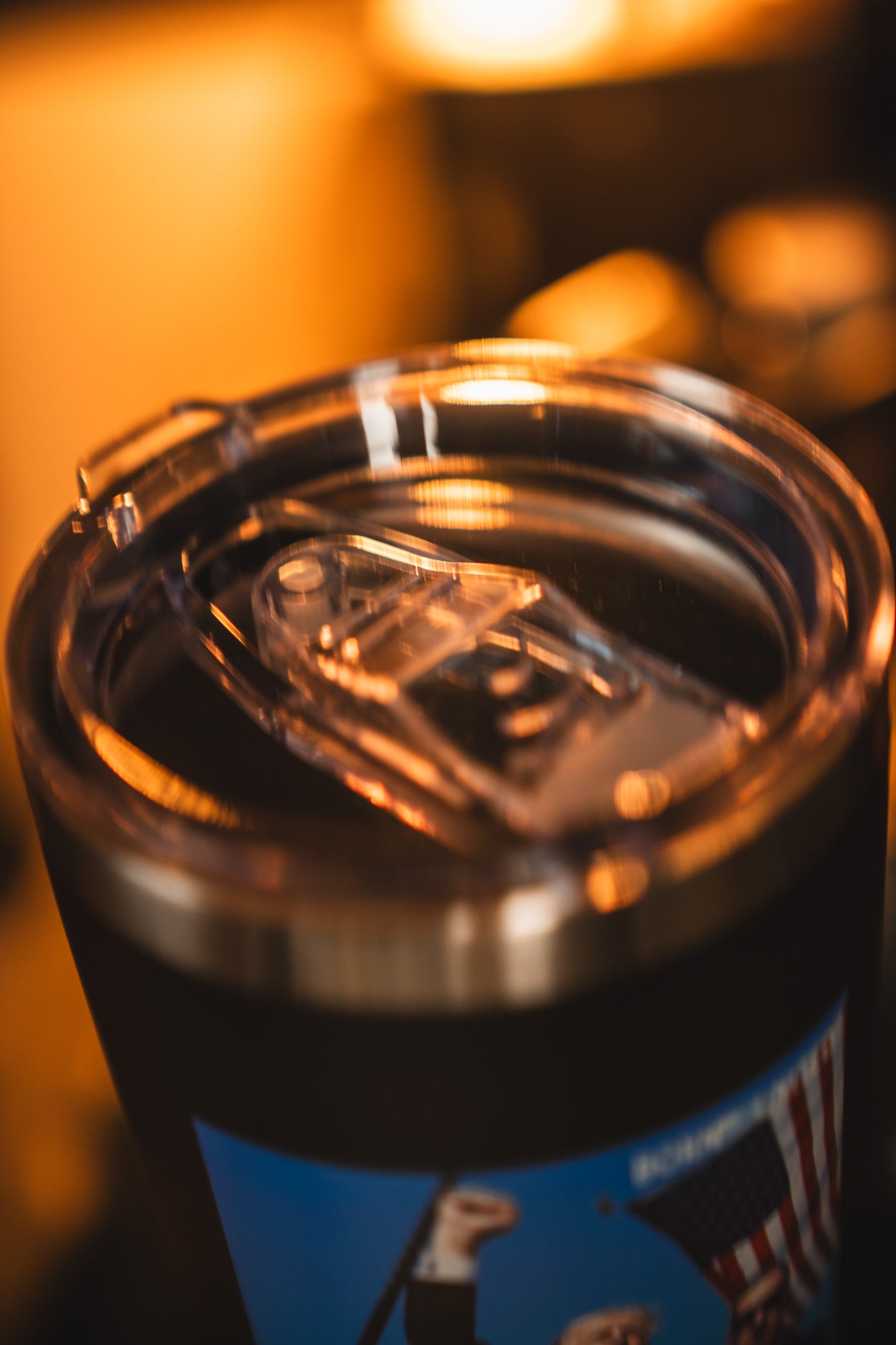 A close-up of the Born United Private Label Forever Legend Tumbler - 20oz in stainless steel with a clear lid, set against a blurred American flag under warm lighting.