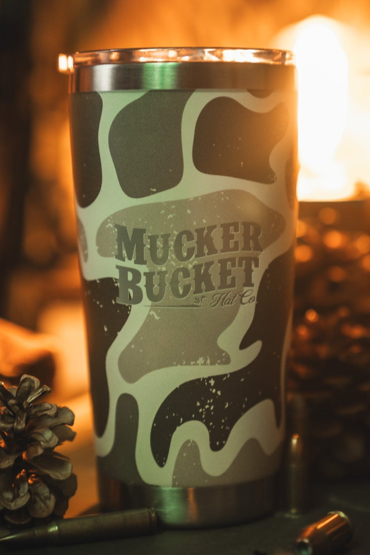 The Mucker Bucket Duck Camo Tumbler - 20oz rests beside pinecones and a bullet casing under warm, blurry light. Its laser-engraved stainless steel design combines outdoor charm with rugged style.