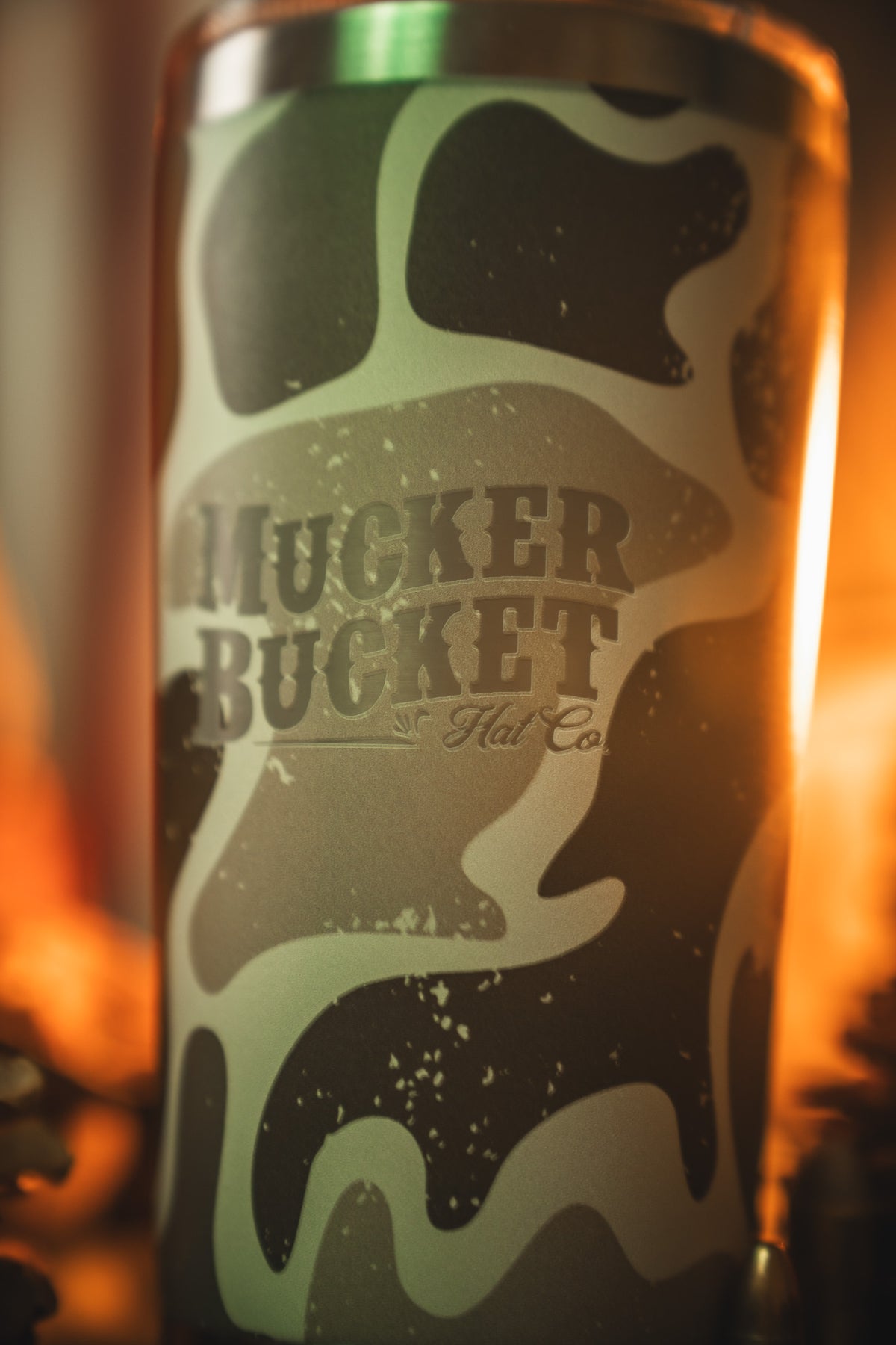A close-up of the Duck Camo Tumbler - 20oz by Mucker Bucket, featuring green and brown camo patterns with the brand logo. Warm lighting adds a cozy touch to this stainless steel tumbler.