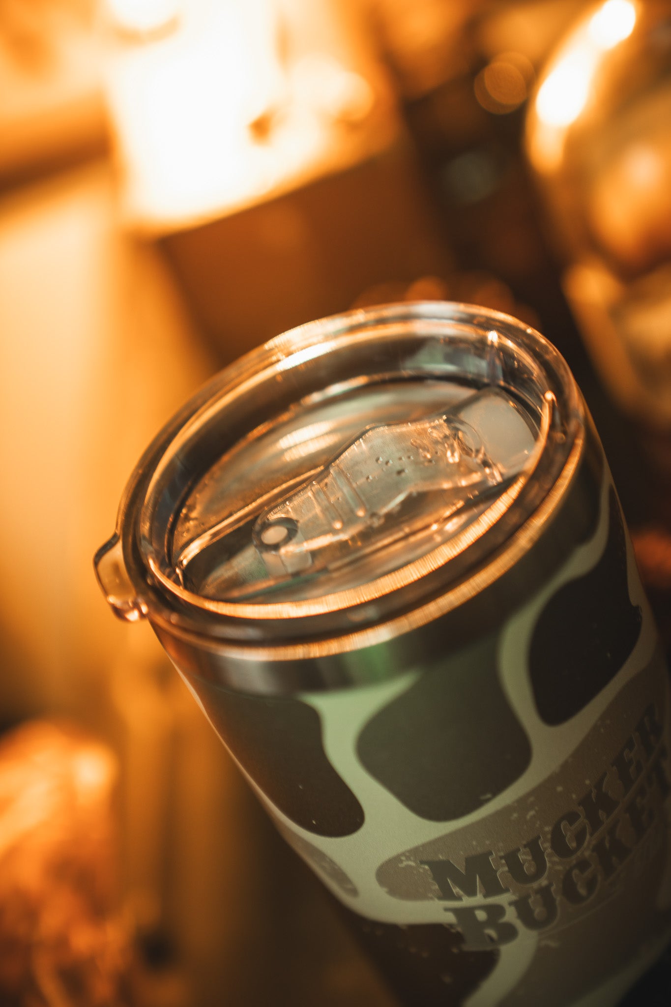 A close-up of the Mucker Bucket Duck Camo Tumbler - 20oz, a stainless steel tumbler with laser engraving and transparent plastic lid, showing a duck camo pattern and "MUCKER BUCKET" text under warm indoor lighting.