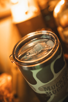 A close-up of the Mucker Bucket Duck Camo Tumbler - 20oz, a stainless steel tumbler with laser engraving and transparent plastic lid, showing a duck camo pattern and "MUCKER BUCKET" text under warm indoor lighting.
