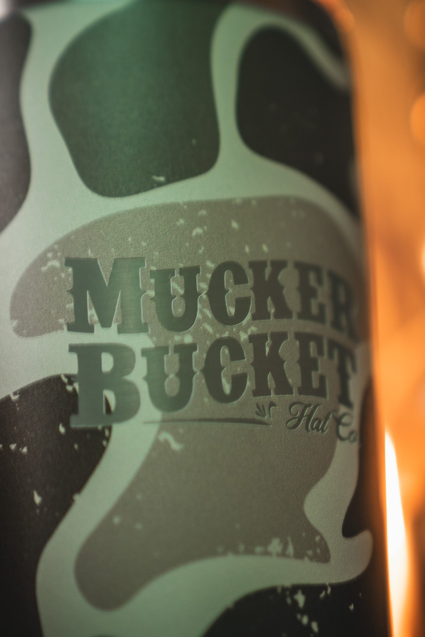 Close-up of the Duck Camo Tumbler - 20oz by Mucker Bucket, featuring the brand's laser-engraved logo on stainless steel. The blurred, warmly lit background enhances its striking green and black camo design.