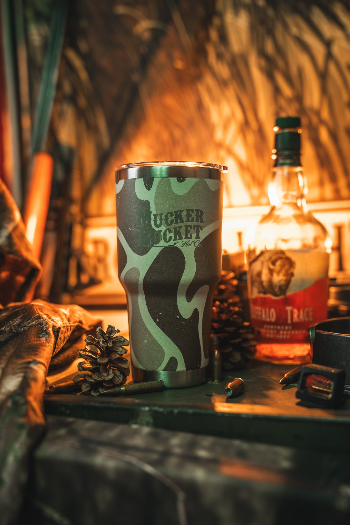 A Mucker Bucket Duck Camo Tumbler - 30oz sits on a table by Buffalo Trace bourbon, pinecones, and bullets, with hunting gear behind. Warm, dramatic light highlights the custom laser-engraved stainless steel tumbler.