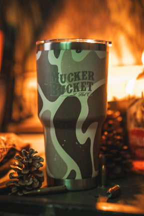The Duck Camo Tumbler - 30oz by Mucker Bucket features a green and tan camouflage design and custom laser engraving, displayed with pinecones, a bullet, and warm ambient lighting.