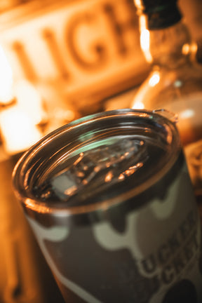 A close-up of the Mucker Bucket Duck Camo Tumbler - 30oz filled with ice and a drink, with a glowing LIGHT sign and bottle blurred in the background—a custom laser-engraved stainless steel tumbler for any setting.