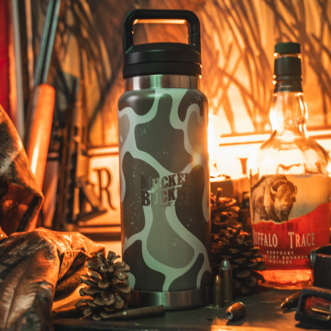 The Mucker Bucket Duck Jug - Duck Camo Water Bottle (26oz) sits amid pinecones, Buffalo Trace whiskey, bullets, and a jacket on a rustic surface. This leak-proof stainless steel bottle is perfect for every adventure.