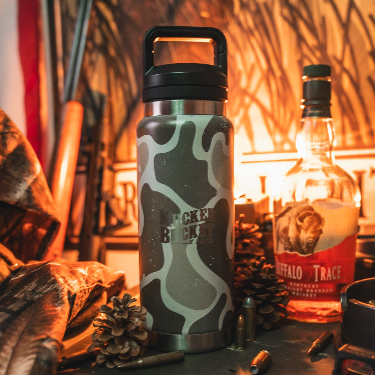 A Mucker Bucket Duck Jug - Duck Camo Water Bottle (26oz) sits on a table with pinecones, bullets, and Buffalo Trace bourbon, all set against a rustic outdoor backdrop.