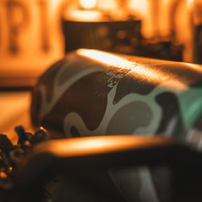 A close-up of the Mucker Bucket Duck Jug - Duck Camo Water Bottle (26oz) highlights its camouflage stainless steel body and leak-proof design, set against a blurred background with warm lighting.