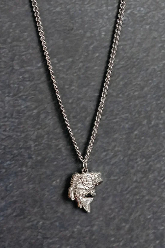 The Big Bass America Necklace by Mucker Bucket is a silver piece featuring a fish-shaped pendant, showcased against a textured dark gray backdrop.