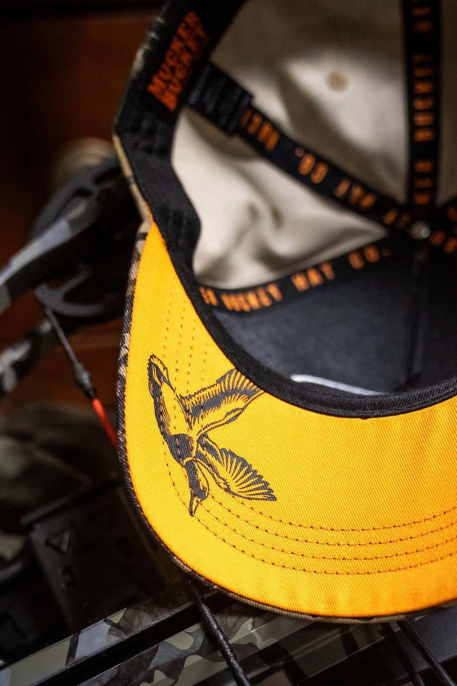 A close-up of the Mucker Bucket Mallard Life Deadwood Camo Bucket hat with a bird design, resting on deadwood camo gear and a crossbow. The hat's interior is beige with black and orange accents.