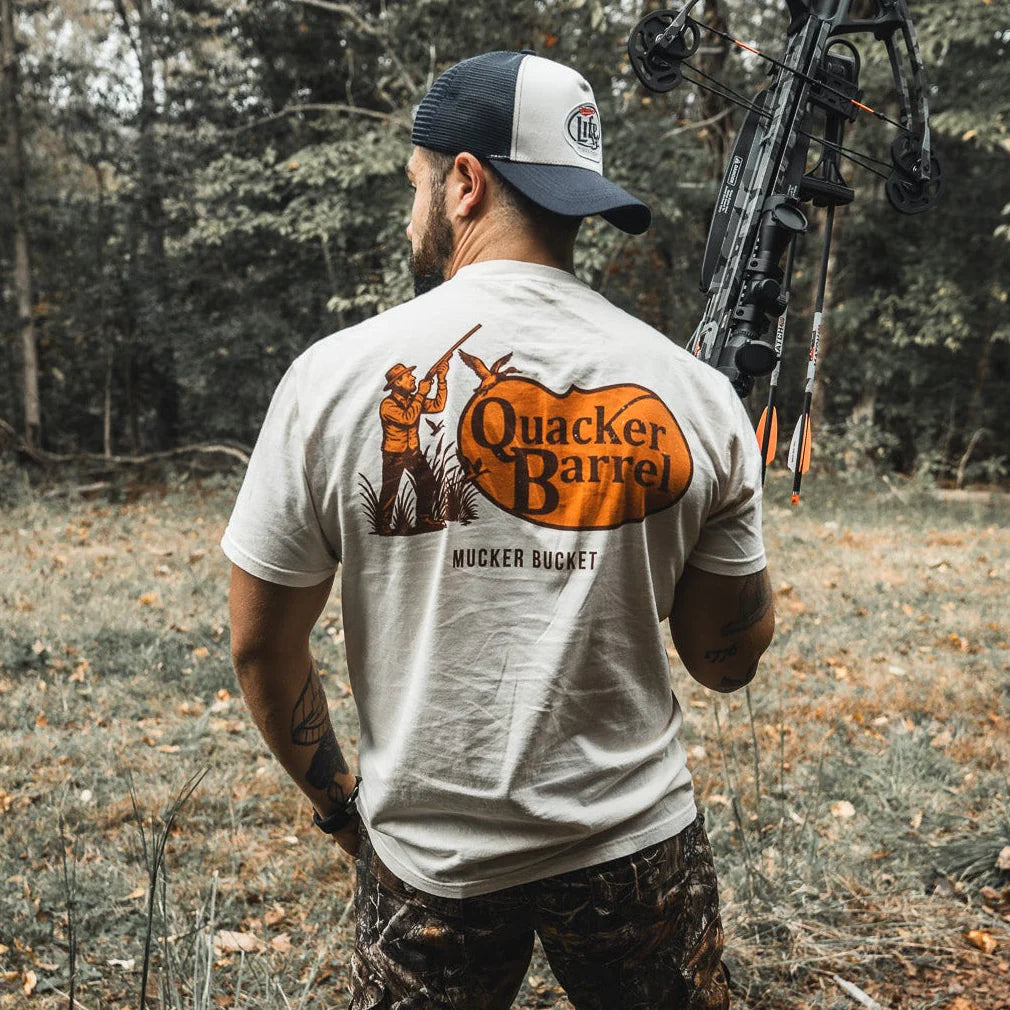 A man in camo pants and a white Mucker Bucket Quacker Barrel Premium Tee stands outdoors with a compound bow, back turned in a leafy wooded area. The premium tee is crafted from soft, garment-dyed cotton for a relaxed fit.