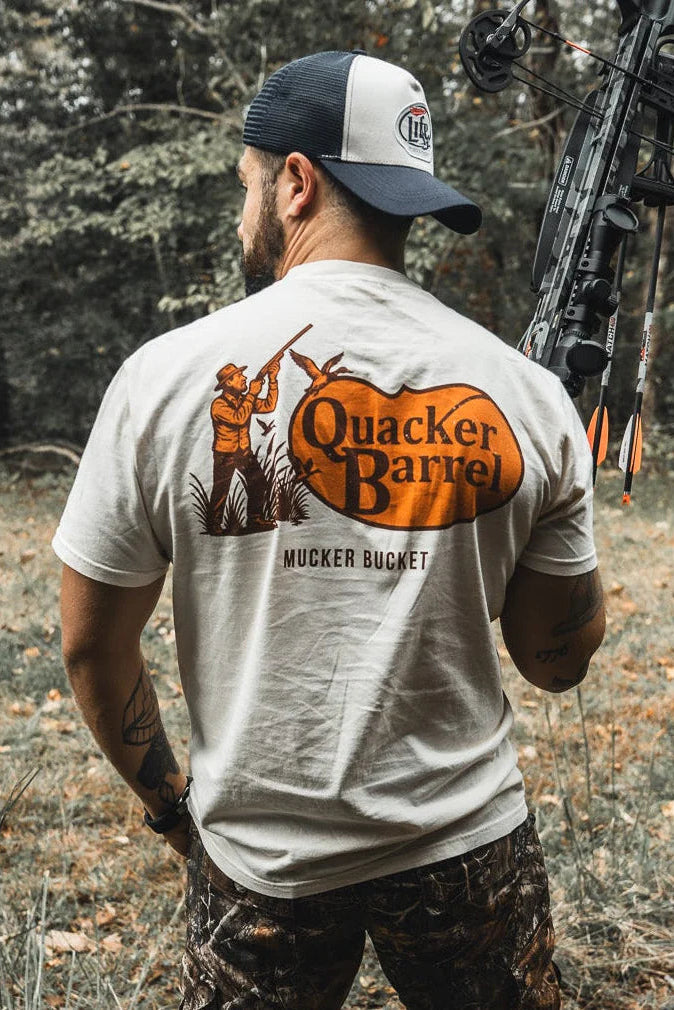 A man in camo pants and a white Mucker Bucket Quacker Barrel Premium Tee stands outdoors with a compound bow, back turned in a leafy wooded area. The premium tee is crafted from soft, garment-dyed cotton for a relaxed fit.